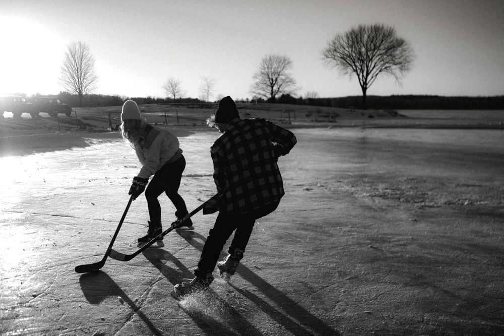 Two people playing hockey on a frozen outdoor pond at sunset in black and white.
