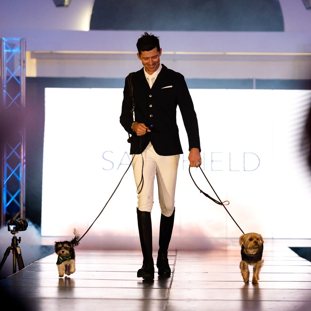 Man in formal attire walking two small dogs on a catwalk or runway.
