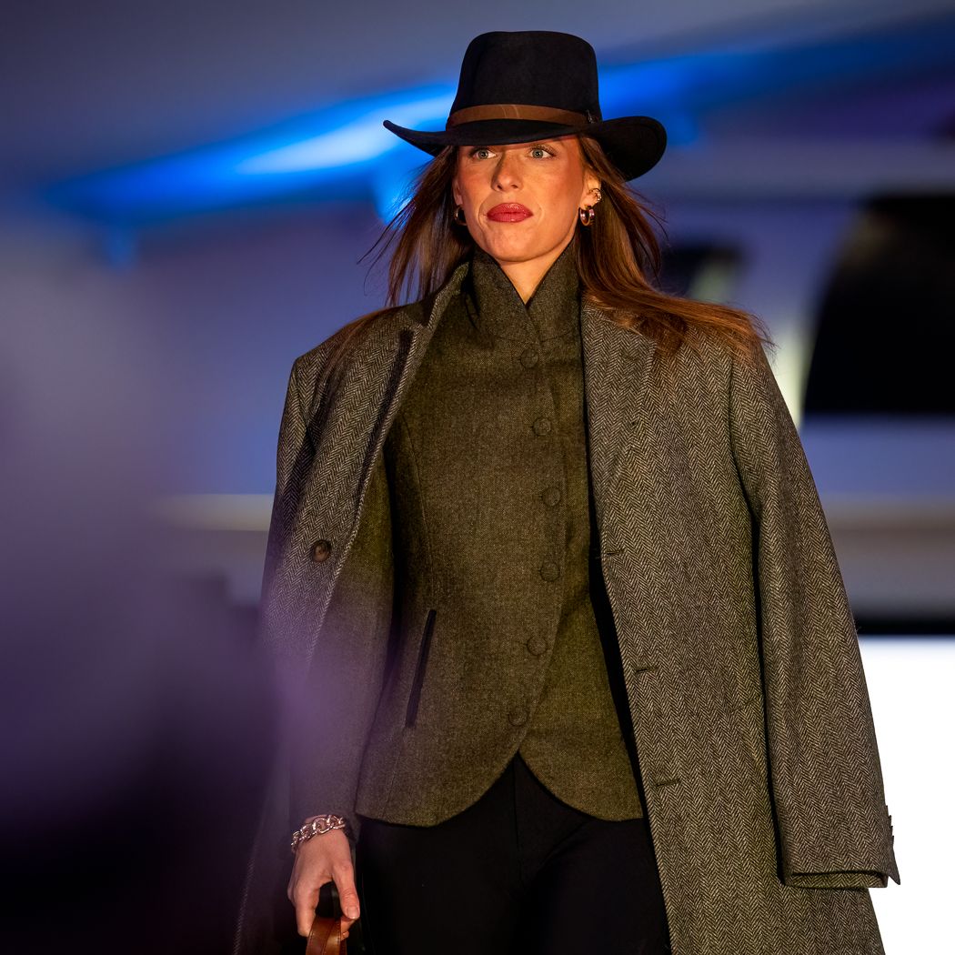 Person on runway wearing a wide-brimmed hat, coat, and dark clothing under blue lighting.