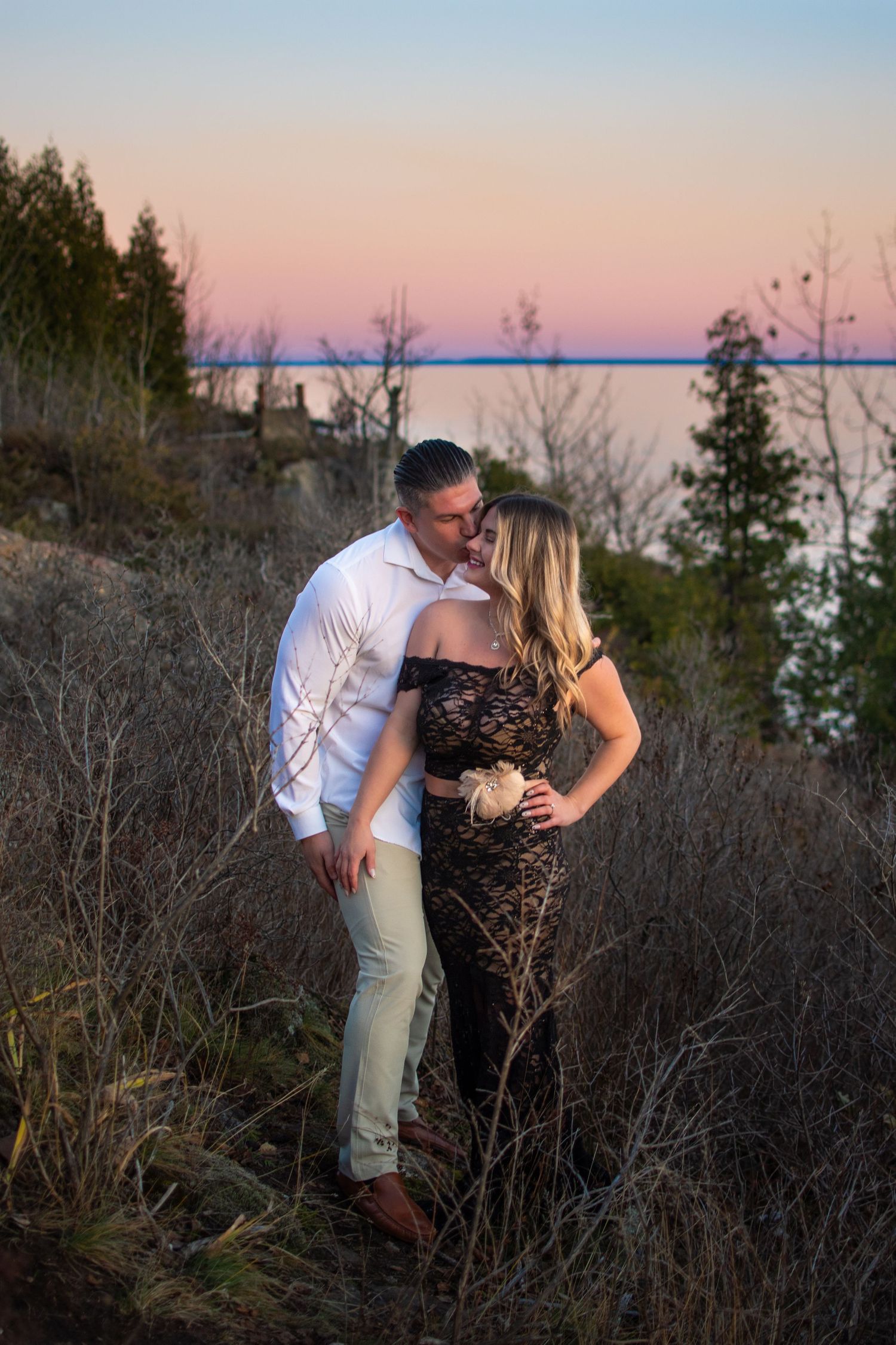 A romantic sunset photo of a couple standing together on a hillside with water in the background.