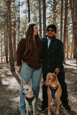 Smiling couple with two dogs standing in sunlit pine forest.