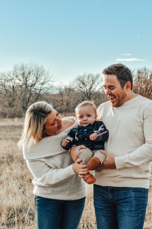 Smiling parents holding adorable baby boy in autumn field under blue sky.