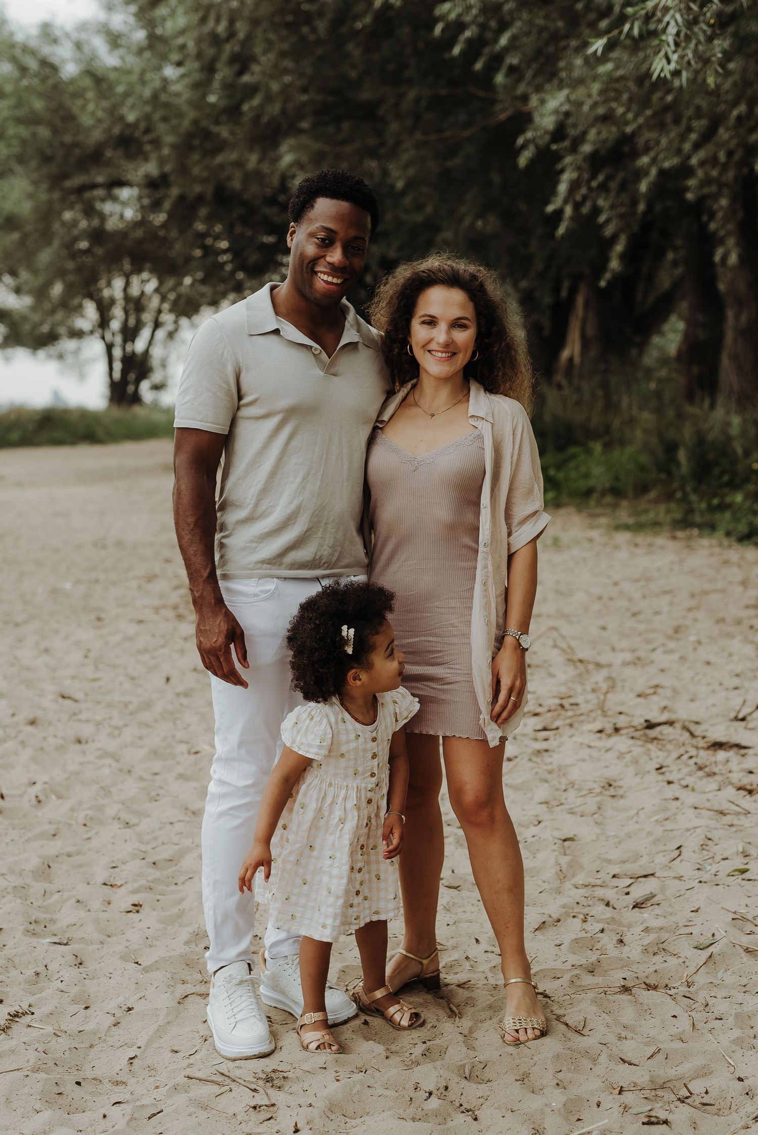 A family of three poses for a portrait on a sandy beach path surrounded by trees.