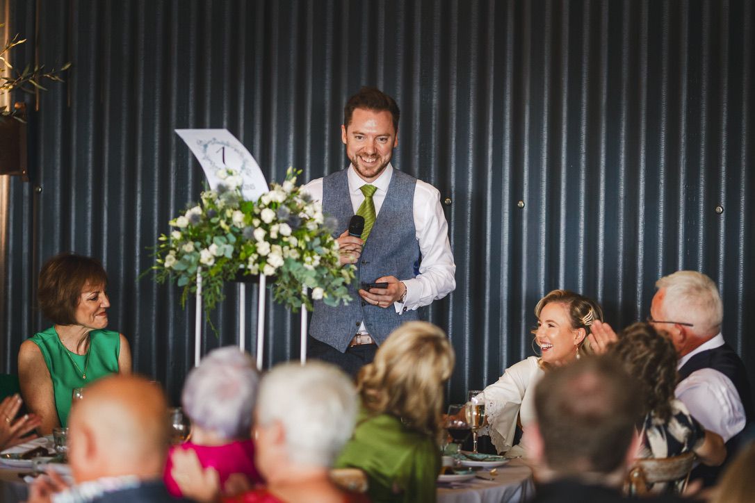 Wedding reception speech being delivered in front of guests at a rustic venue.