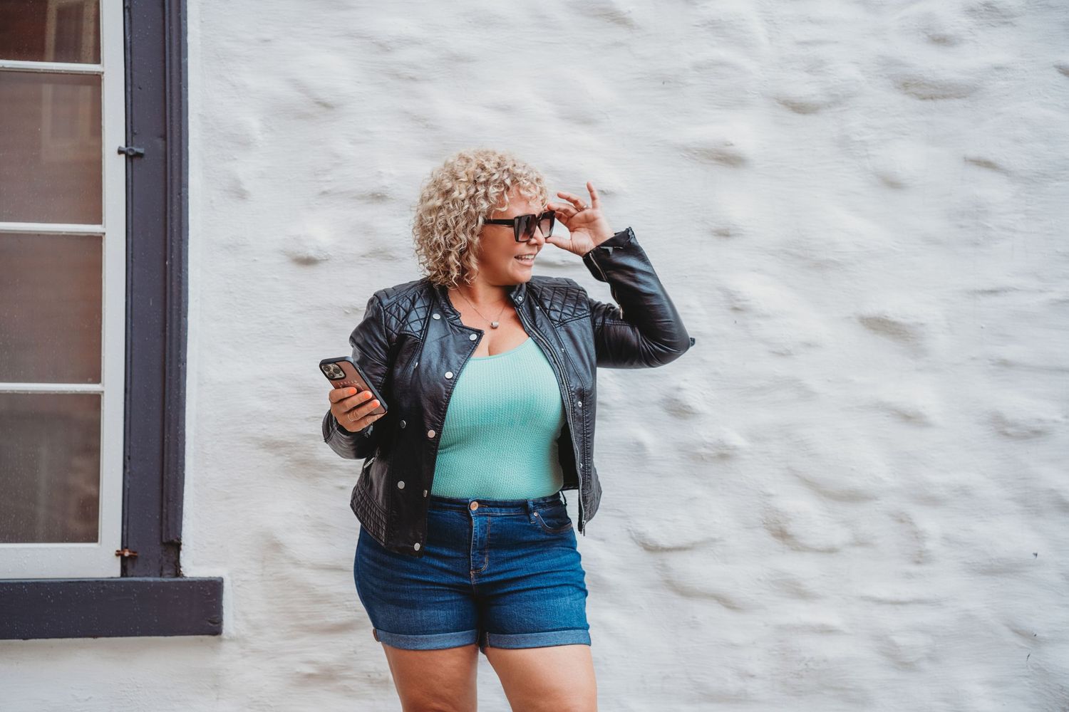 Woman in leather jacket and denim shorts poses against white wall while checking phone.