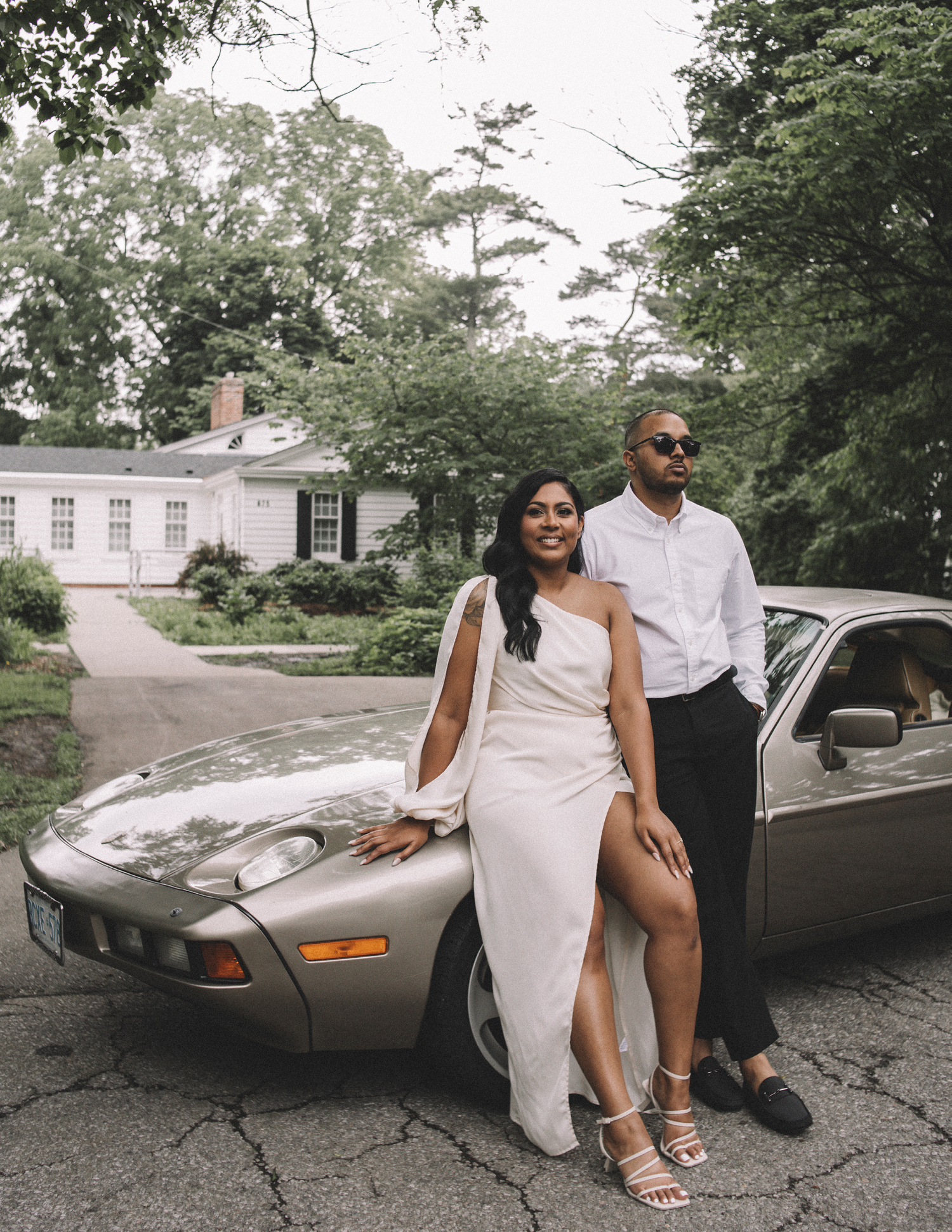 Stylish couple posing with vintage sports car in front of white mansion.