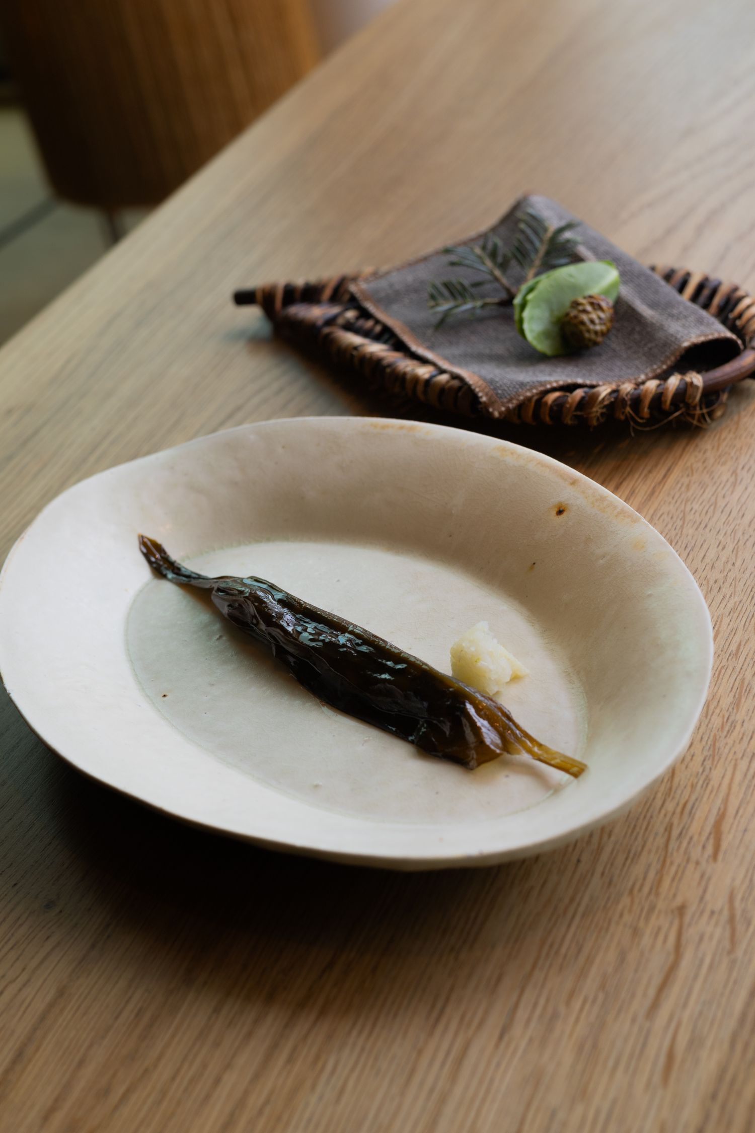 A roasted dark fish served on a simple white plate with a woven basket in the background.