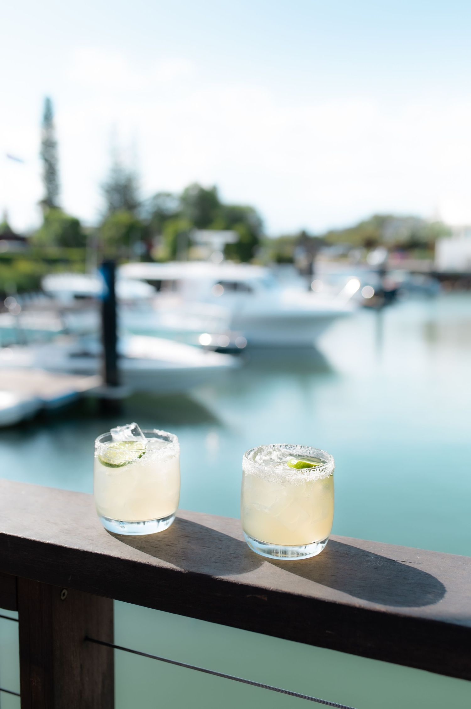 Two cocktail glasses on a railing overlooking a marina with boats in the background.