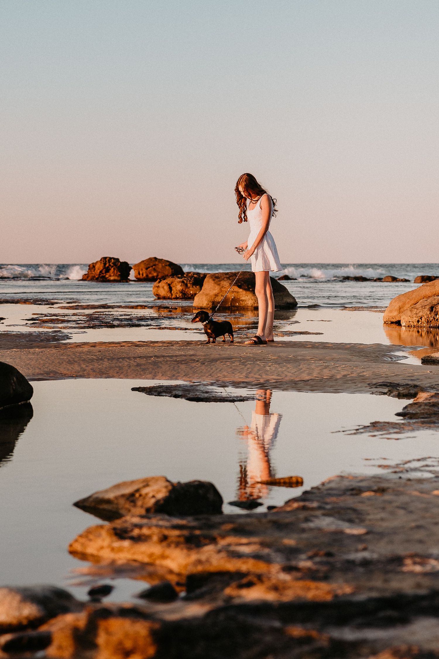 A person in a white dress walking on rocky beach during sunset with reflections in tidal pools.