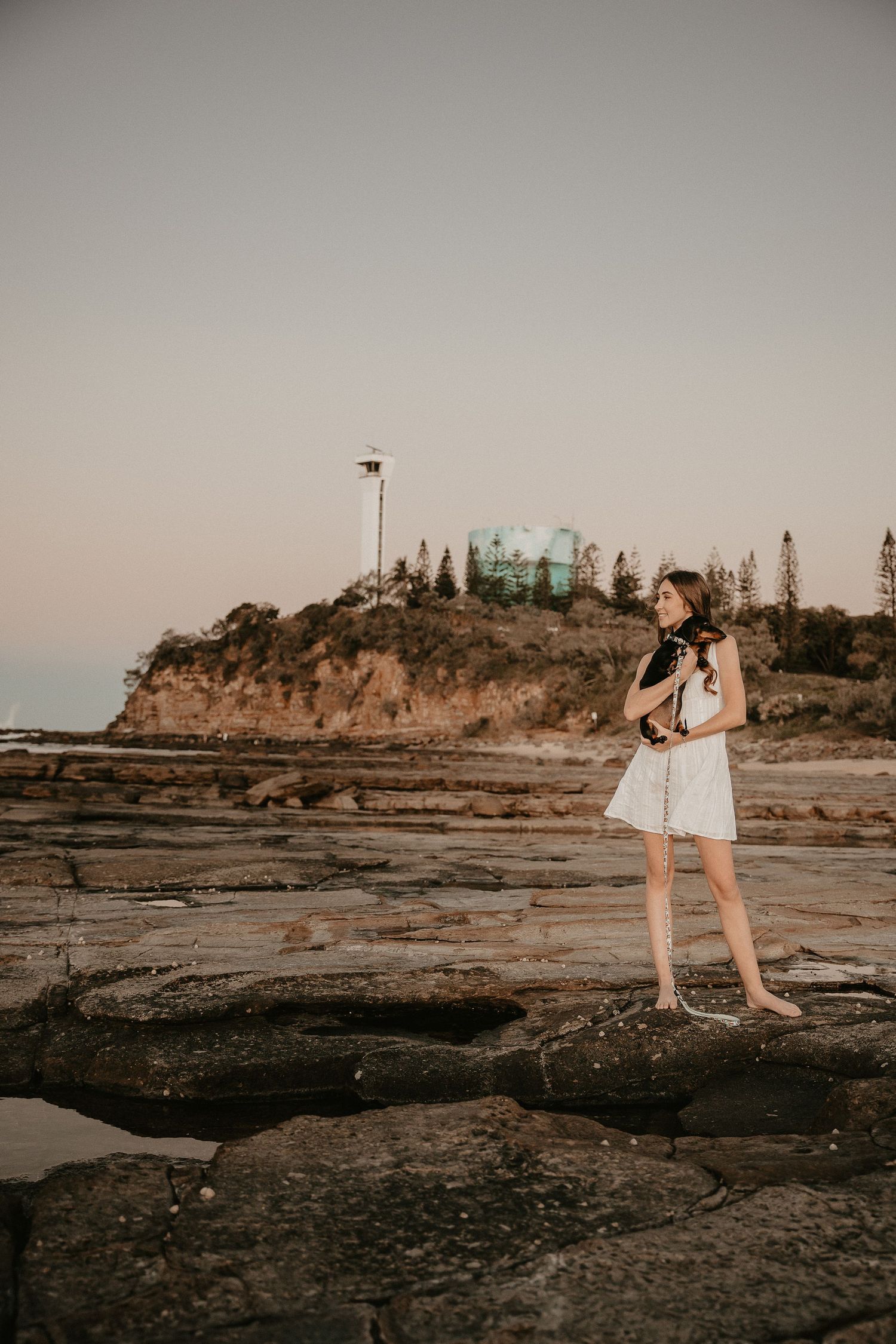 Person in white dress stands on rocky coastal outcrop during sunset.