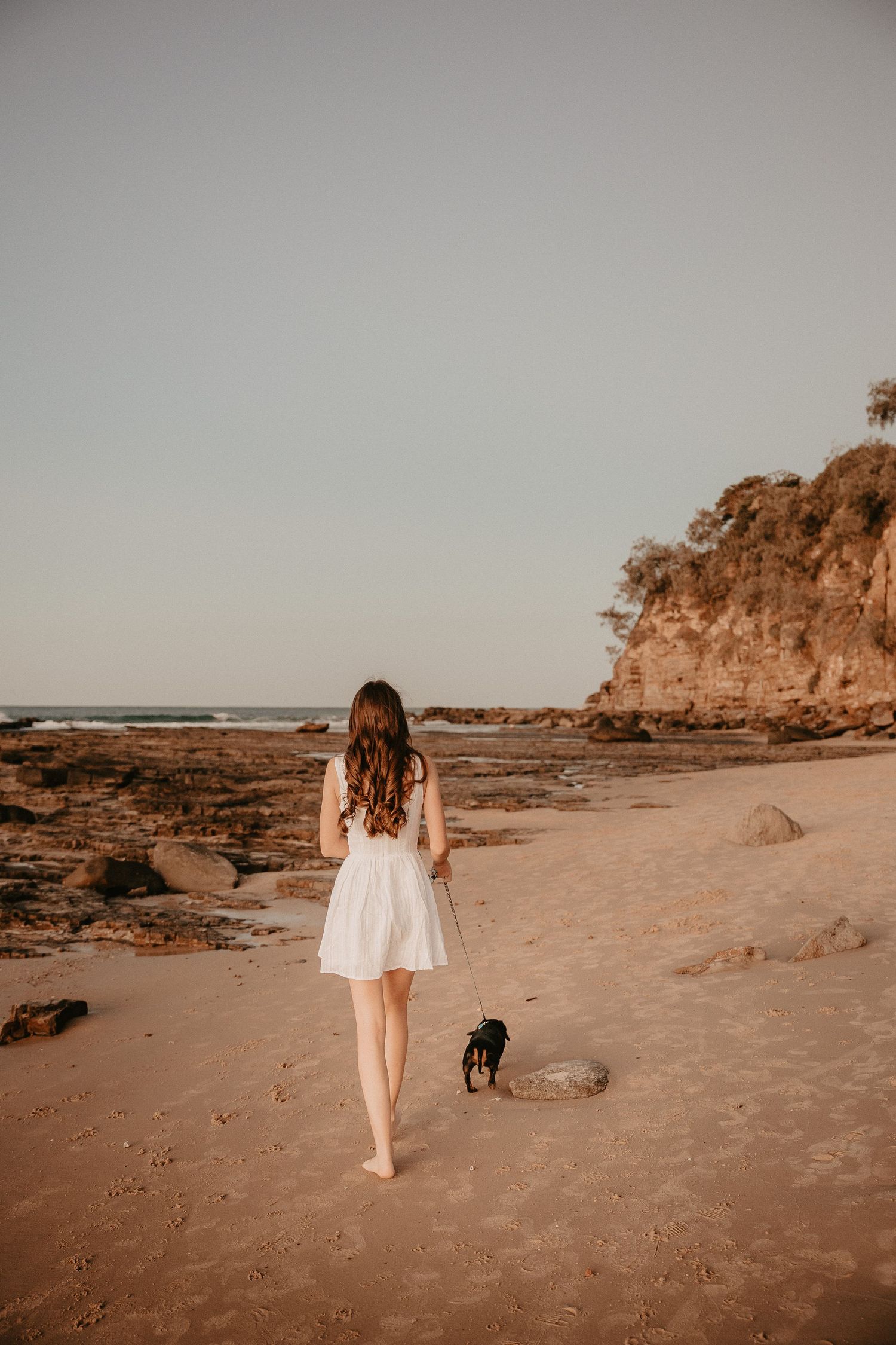 Woman in white dress walking barefoot on beach with small dog following behind.