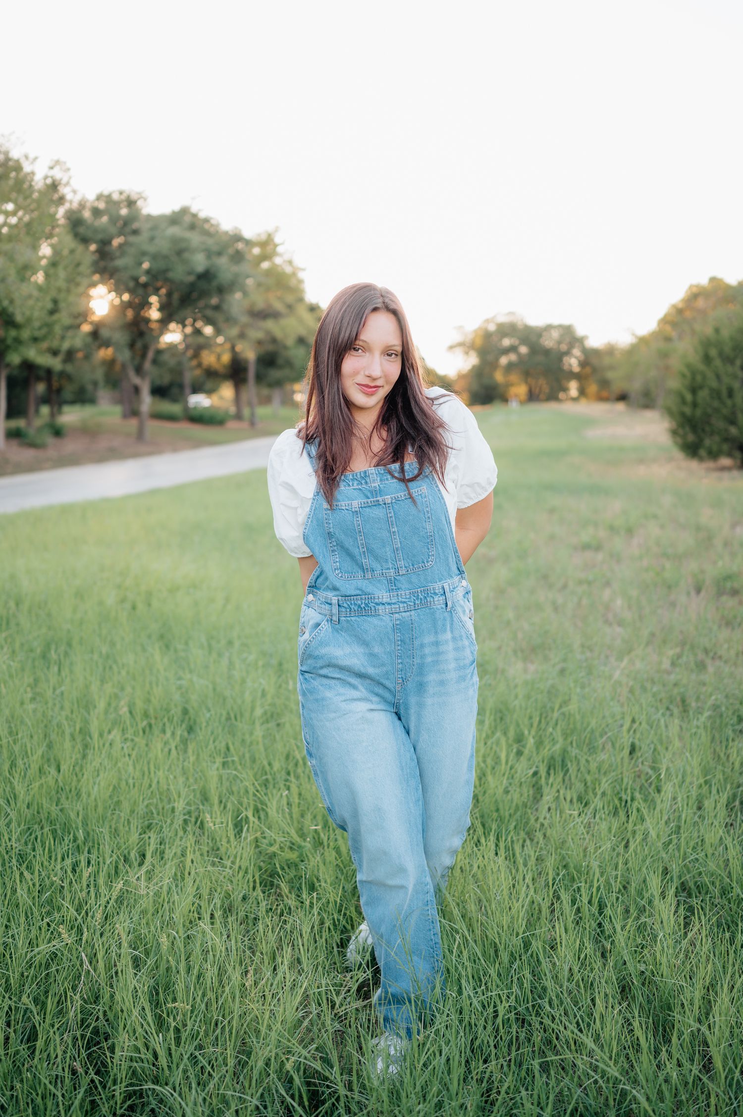 Person in denim overalls and white top walking through tall grass at sunset.