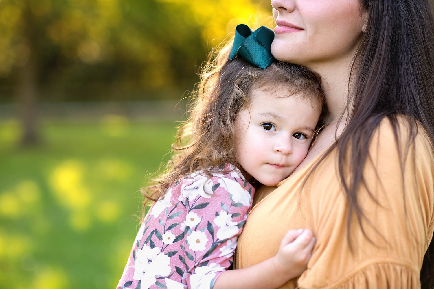 A mother tenderly holds her young daughter with a teal bow in a sunny outdoor setting.