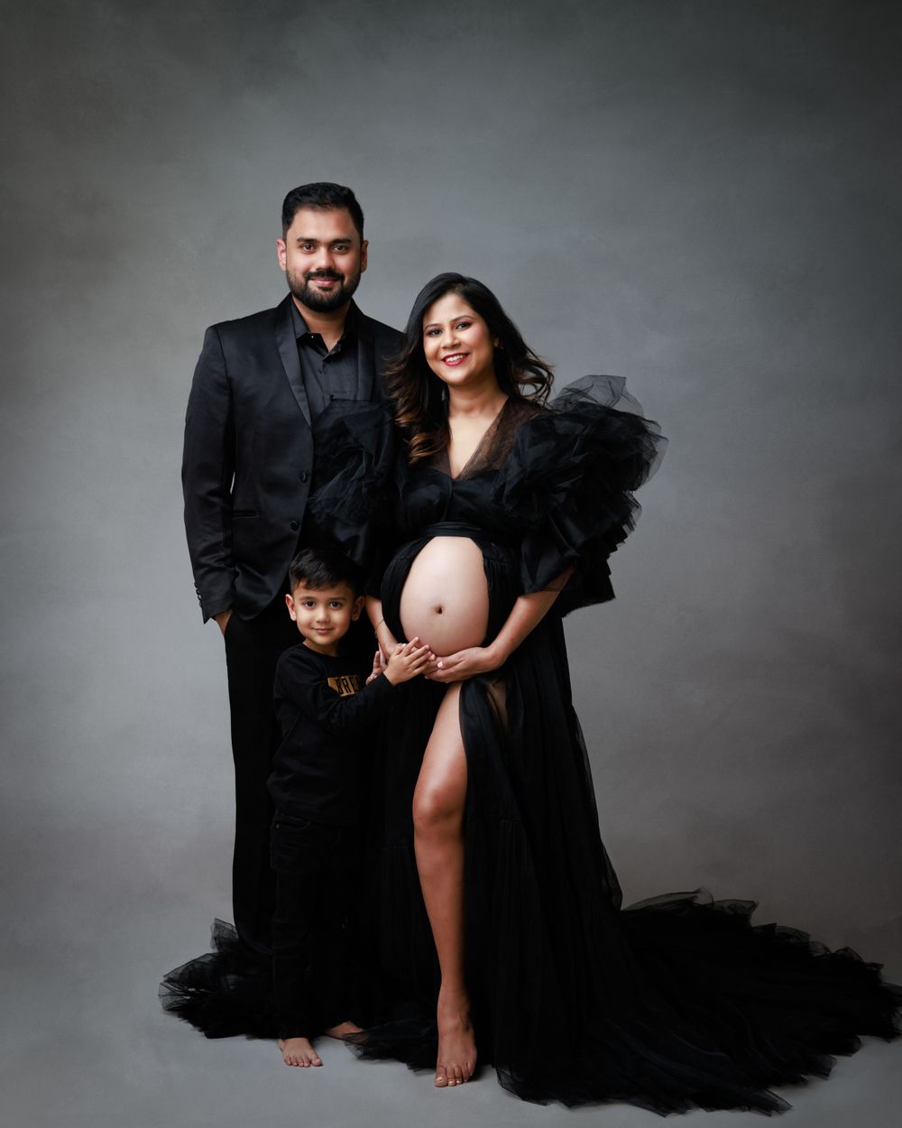 A family maternity portrait with everyone dressed in elegant black attire against a grey background.
