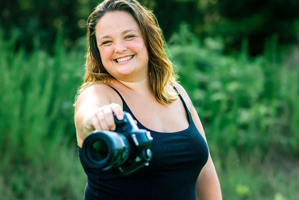 Photographer with long hair smiles while pointing camera at viewer in outdoor nature setting.