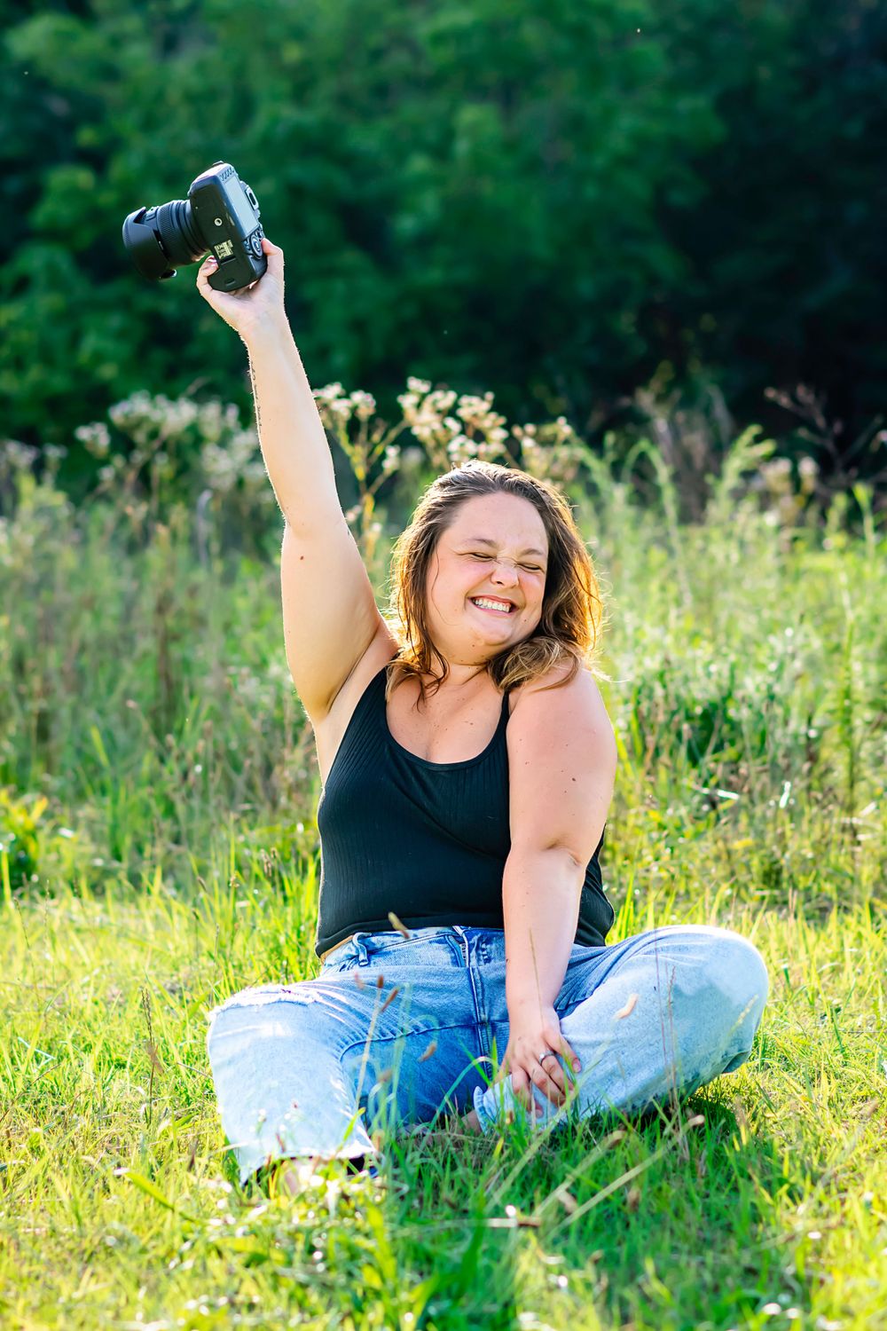 A photographer sits in a grassy field holding a camera up while smiling in the sunlight.