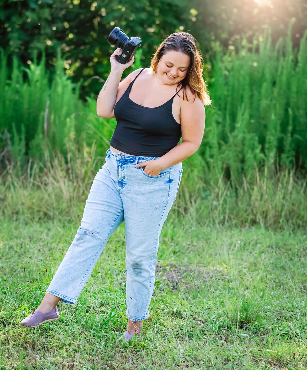 Happy photographer posing playfully in casual outfit with camera against lush green background.
