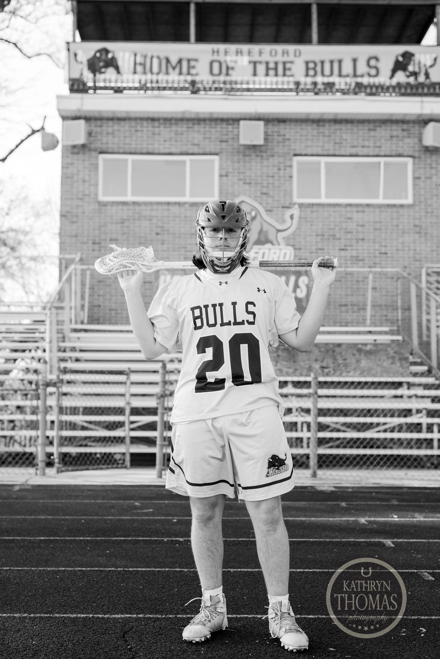 Black and white photo of a player in Bulls number 20 uniform holding lacrosse sticks in front of a stadium.