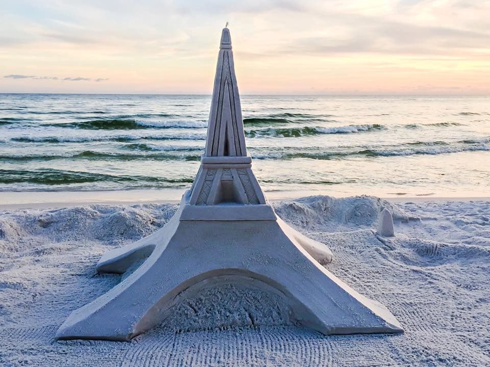 An intricate sand sculpture of the Eiffel Tower on a beach at sunset with waves in the background.