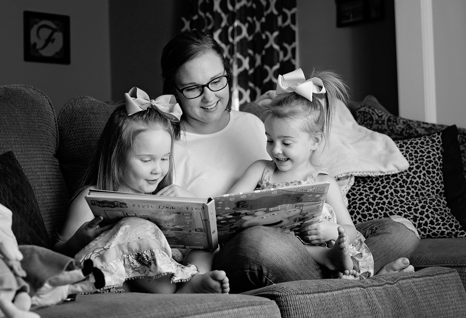 A mother reads a colorful picture book with her two daughters on a cozy sofa in black and white.