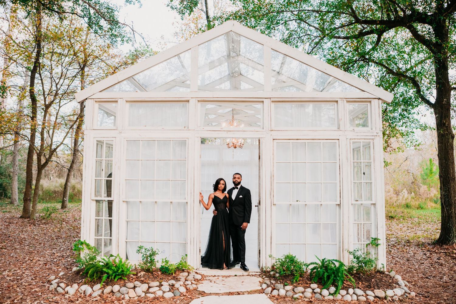 A couple stands under an elegant white greenhouse structure surrounded by trees and fallen leaves in a romantic setting.