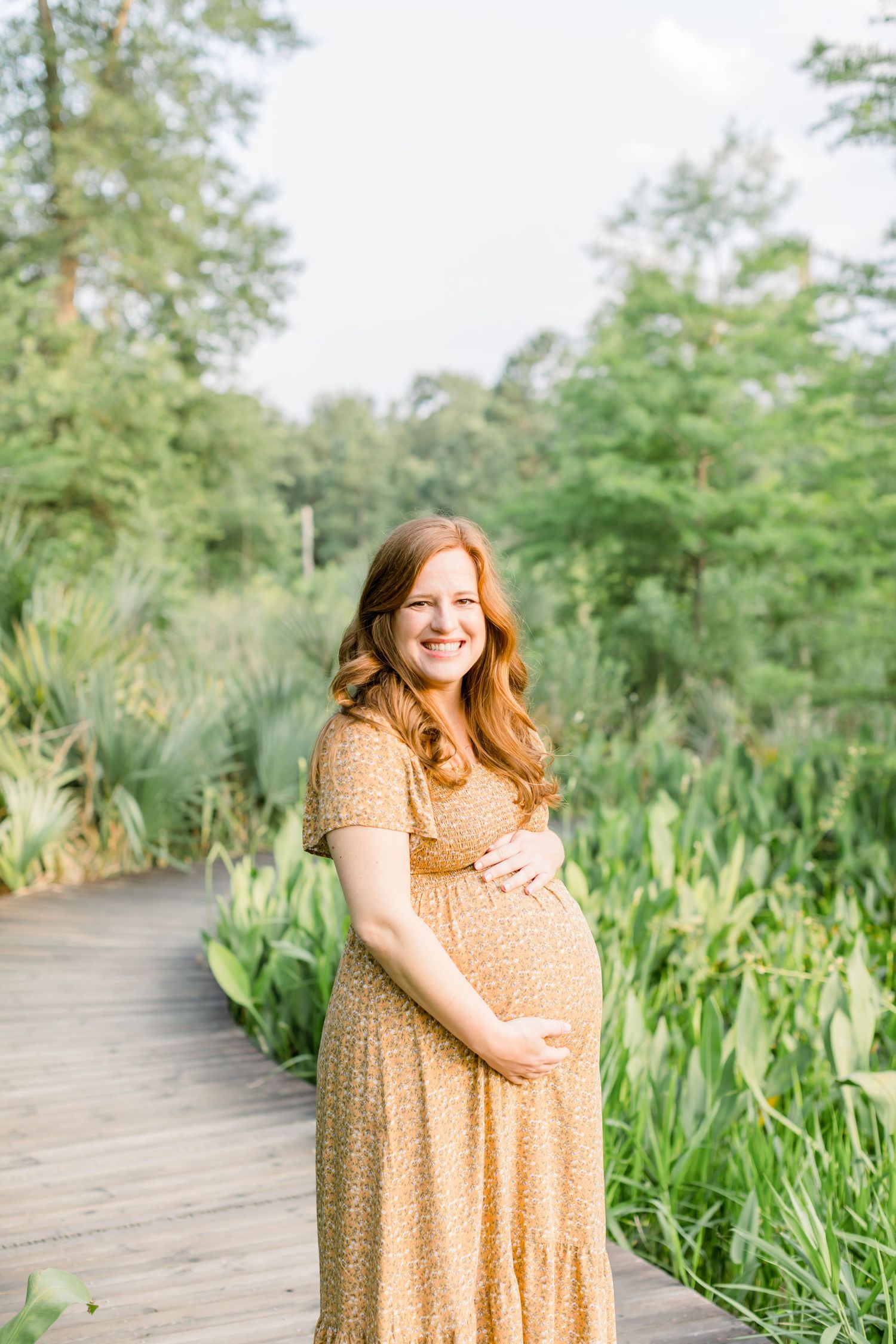 Expectant mother in peach floral dress poses on wooden boardwalk surrounded by lush greenery.