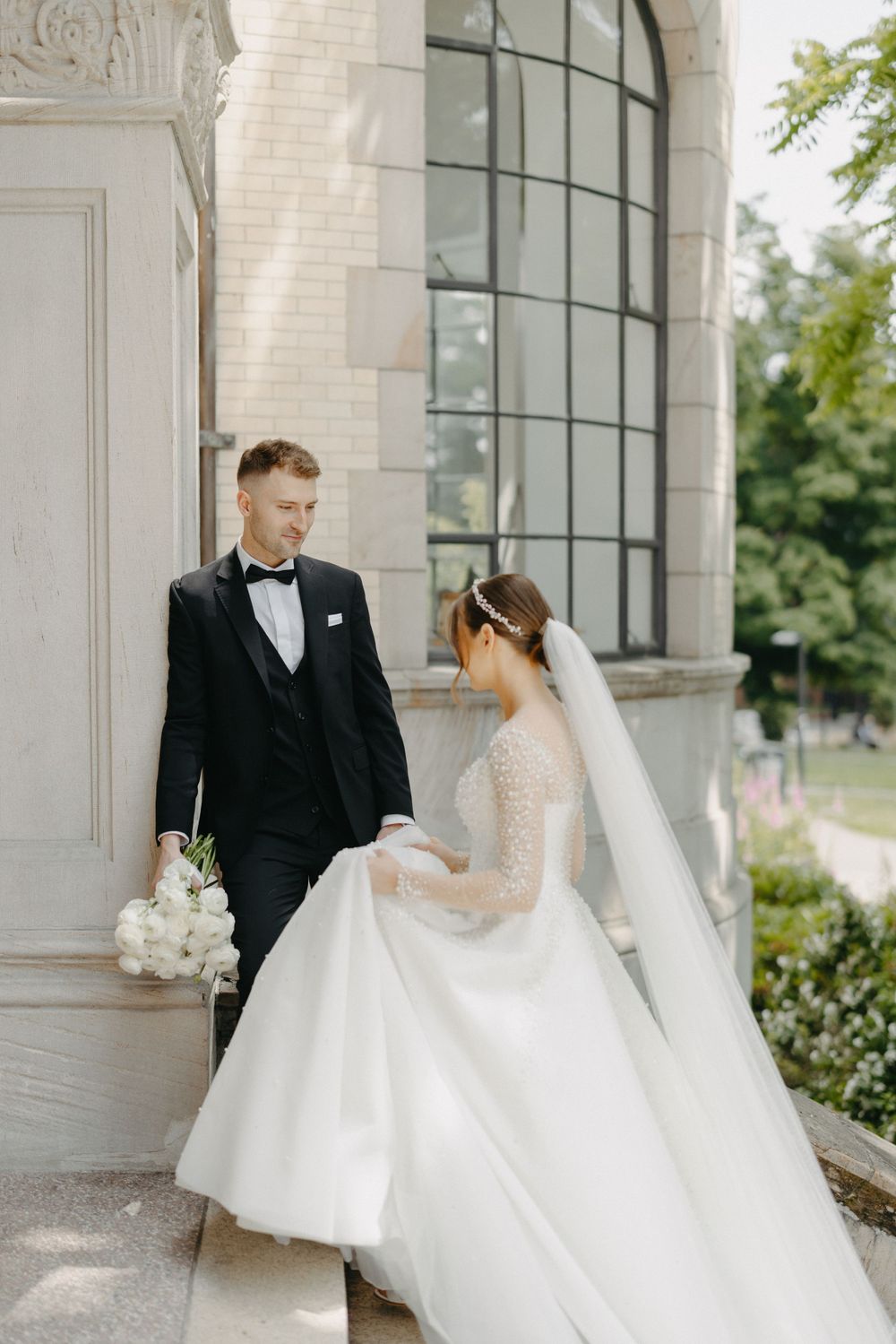 Elegant couple in formal wedding attire sharing a tender moment outside a grand building with arched windows.