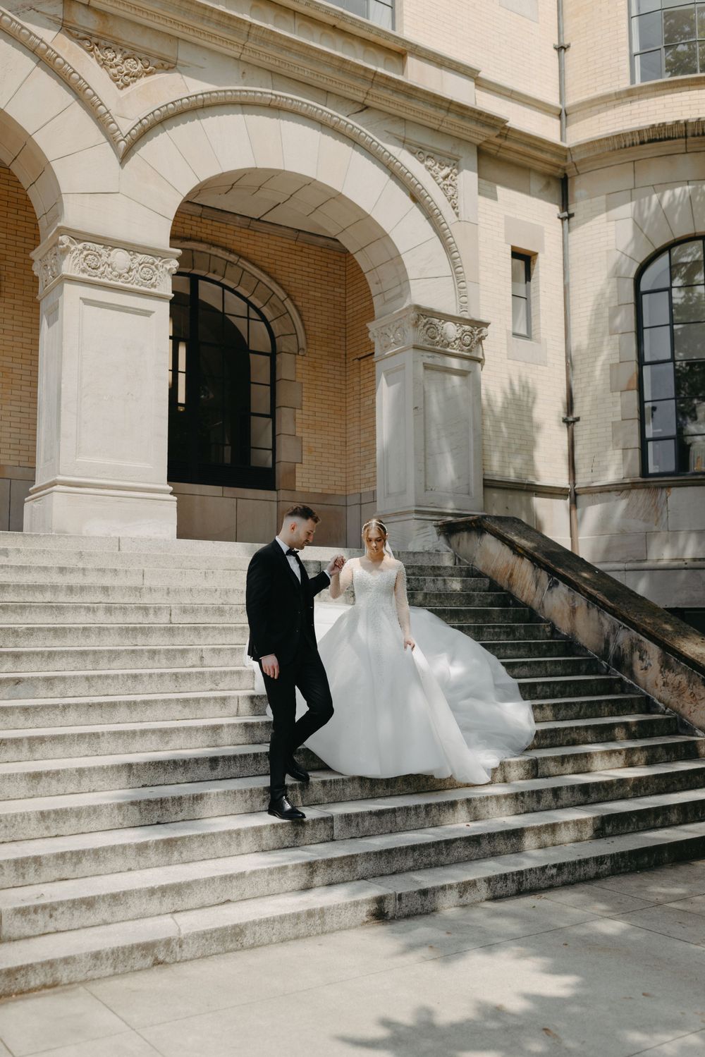 Wedding couple descending grand stone steps of a historic building with arched architecture.