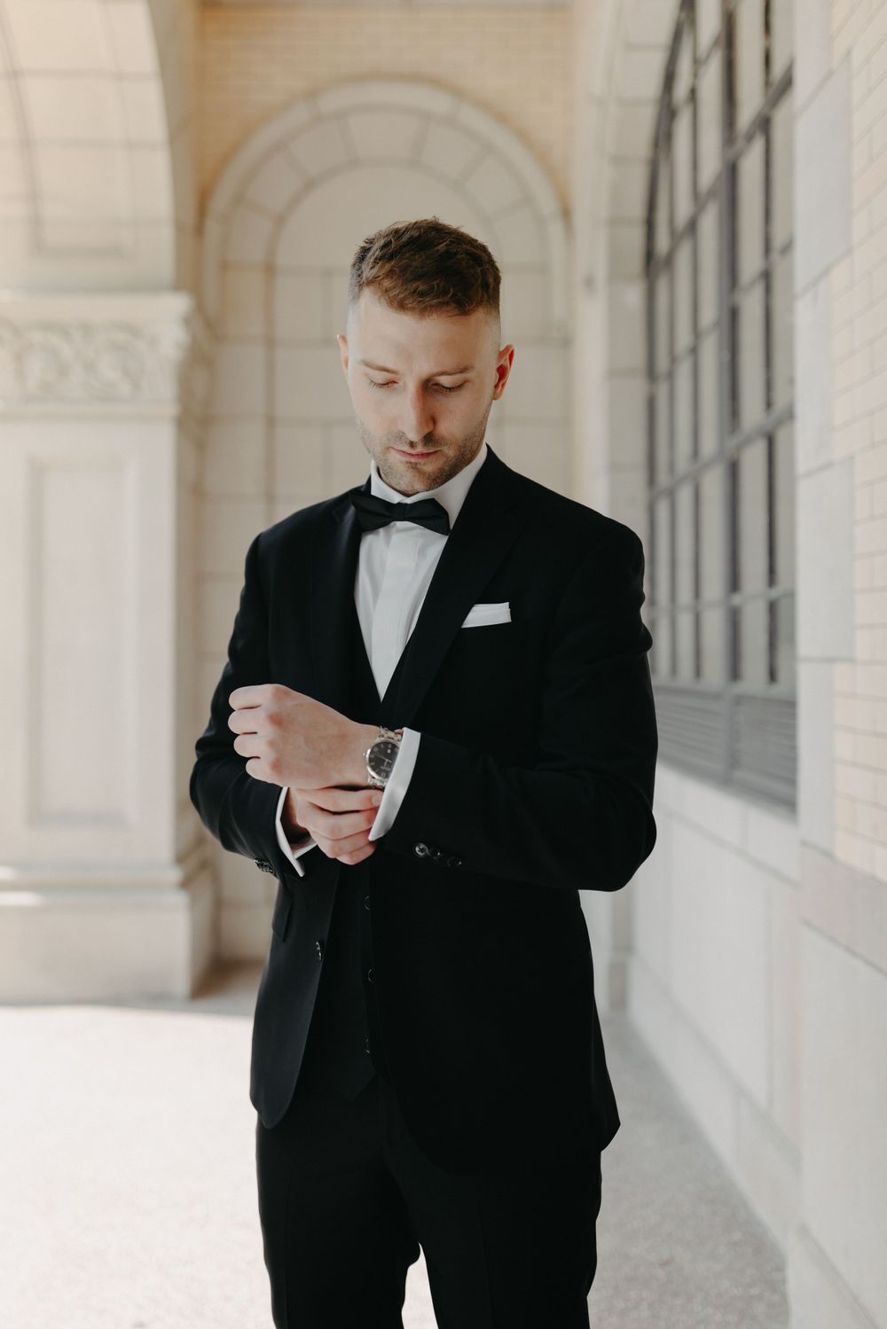 A groom in classic black tuxedo adjusts his cufflinks in an elegant archway.