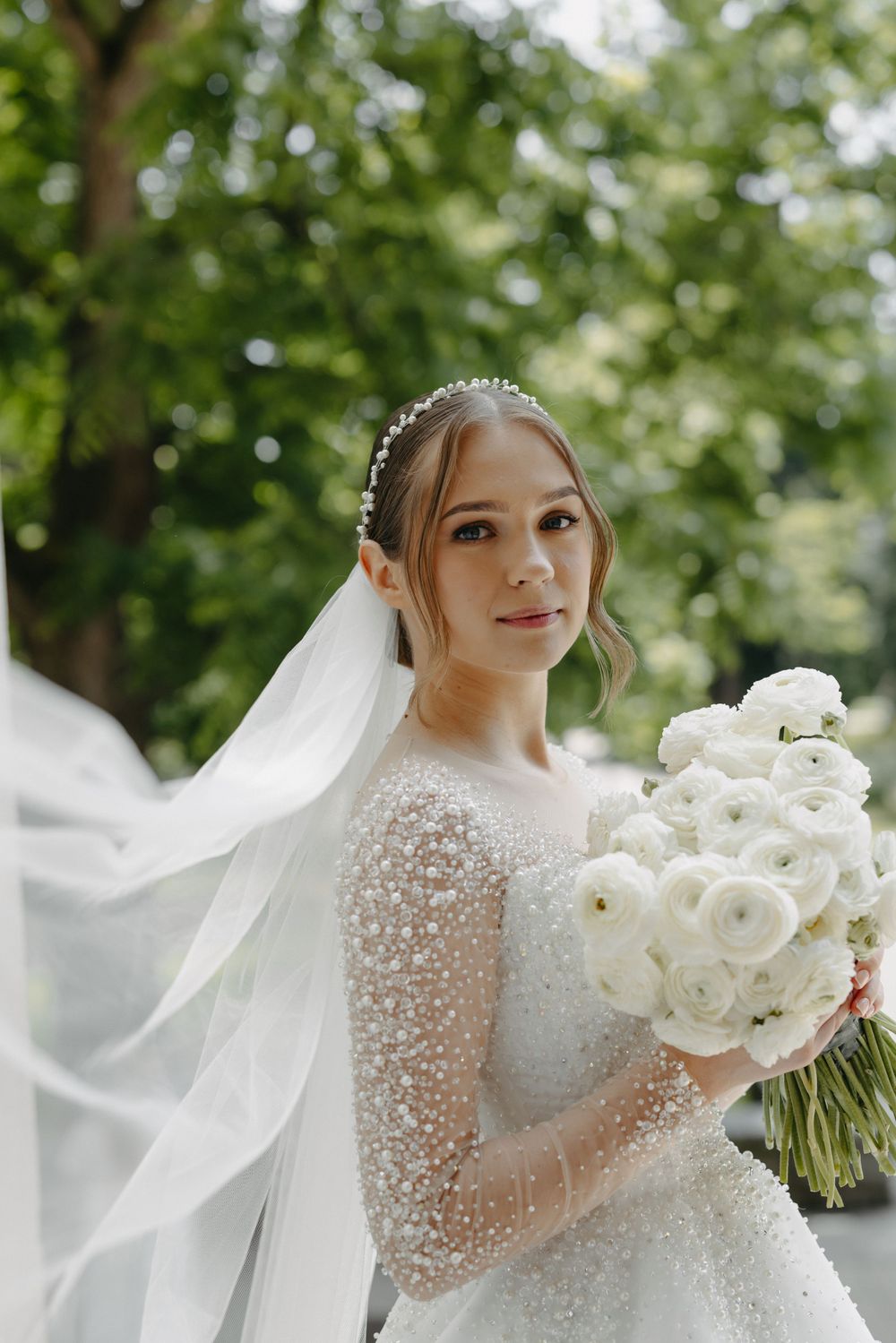 Bride in sparkling long-sleeve wedding gown holds white rose bouquet against natural garden backdrop.