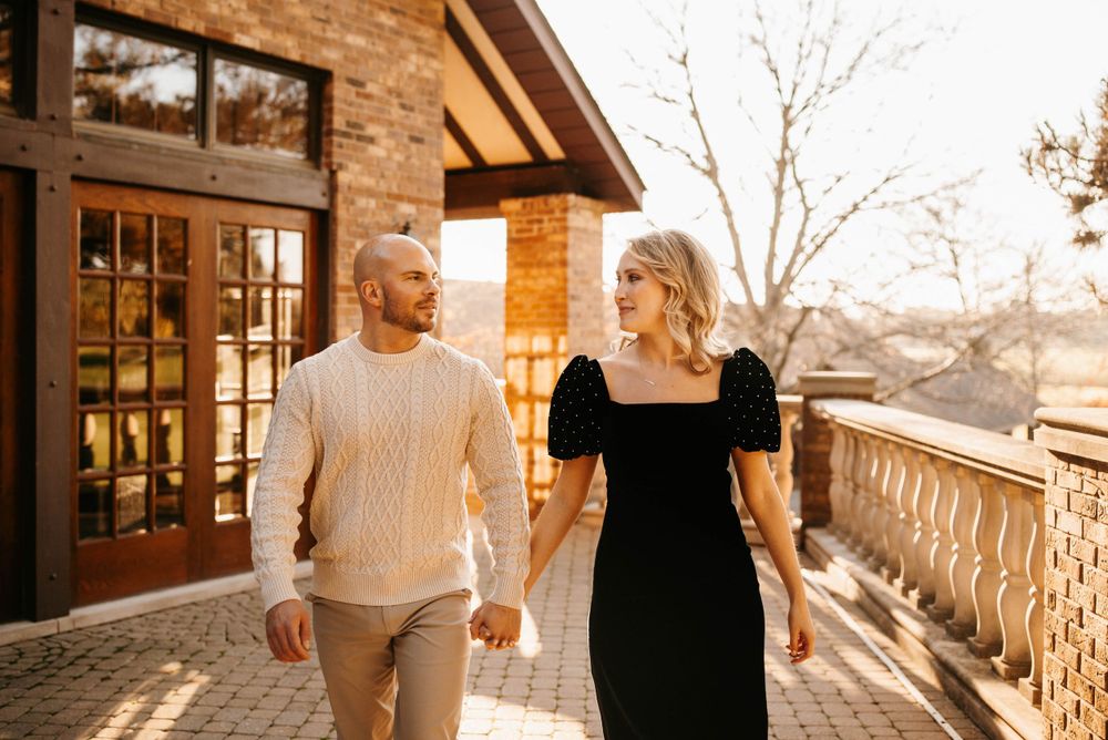 Couple walks hand in hand at golden hour outside a stone building, woman in black dress, man in white sweater.