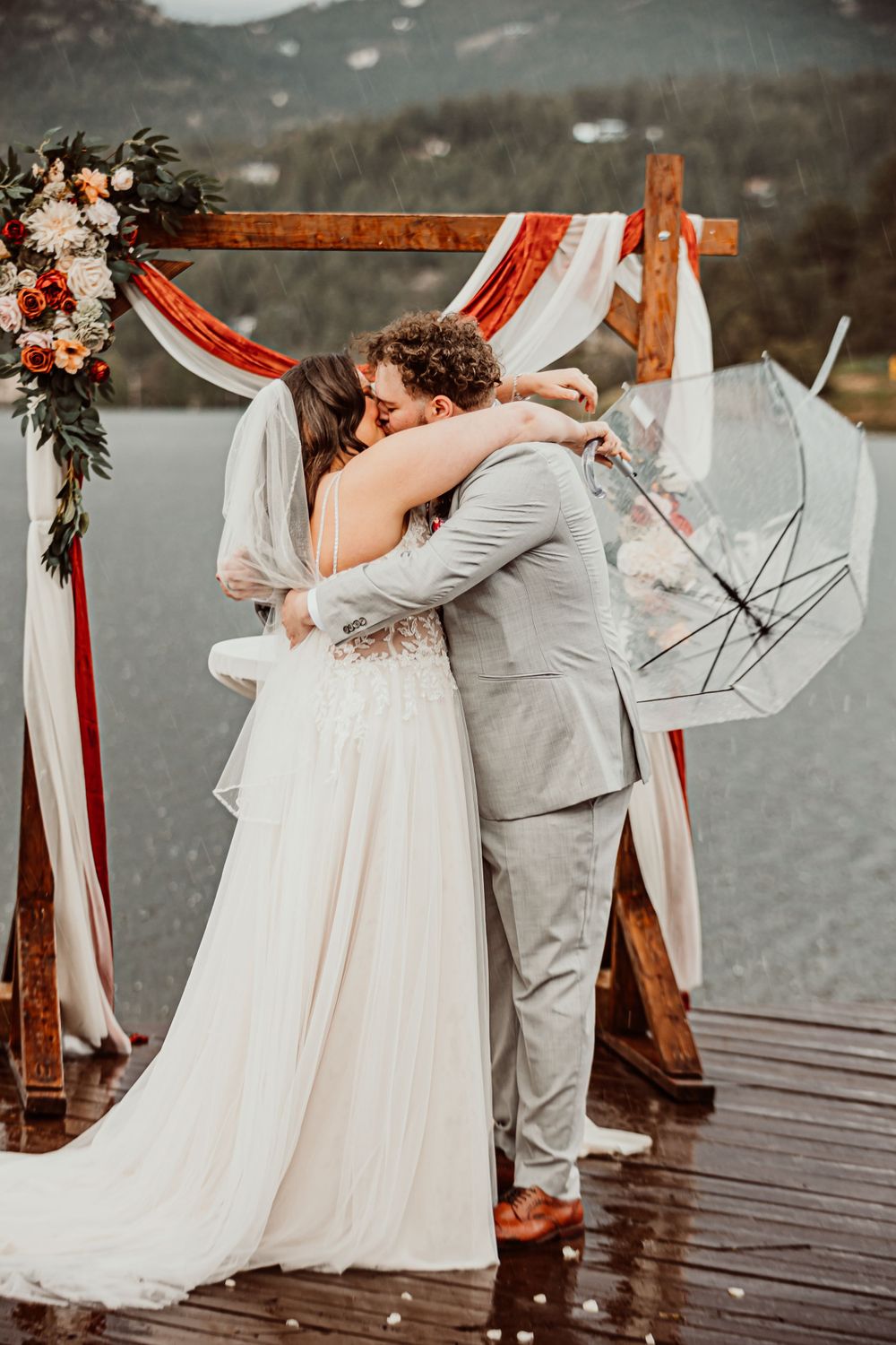 A couple embraces under a floral arch during a rainy mountain wedding ceremony.