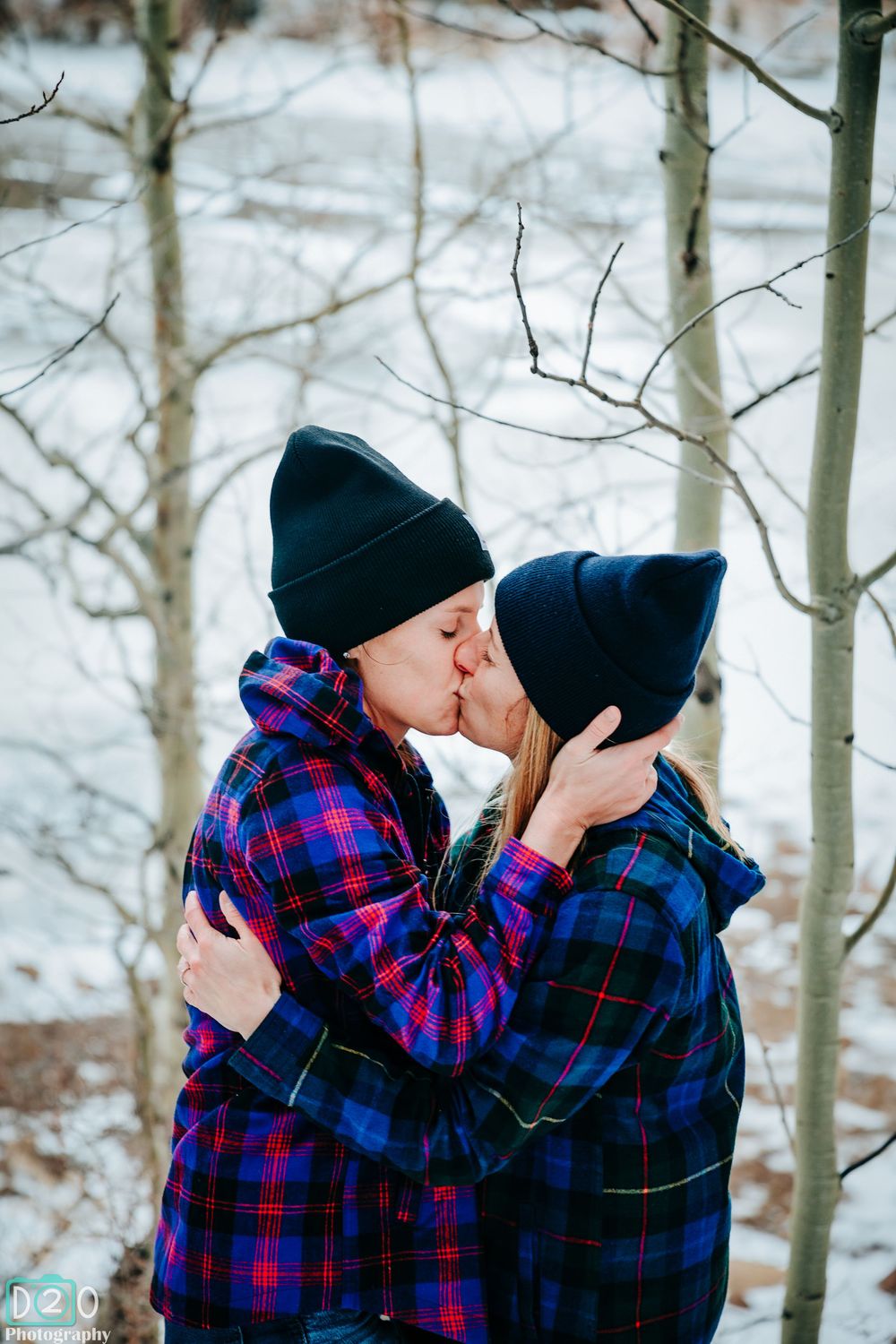 Couple in matching plaid shirts and beanies sharing a kiss in snowy winter forest.