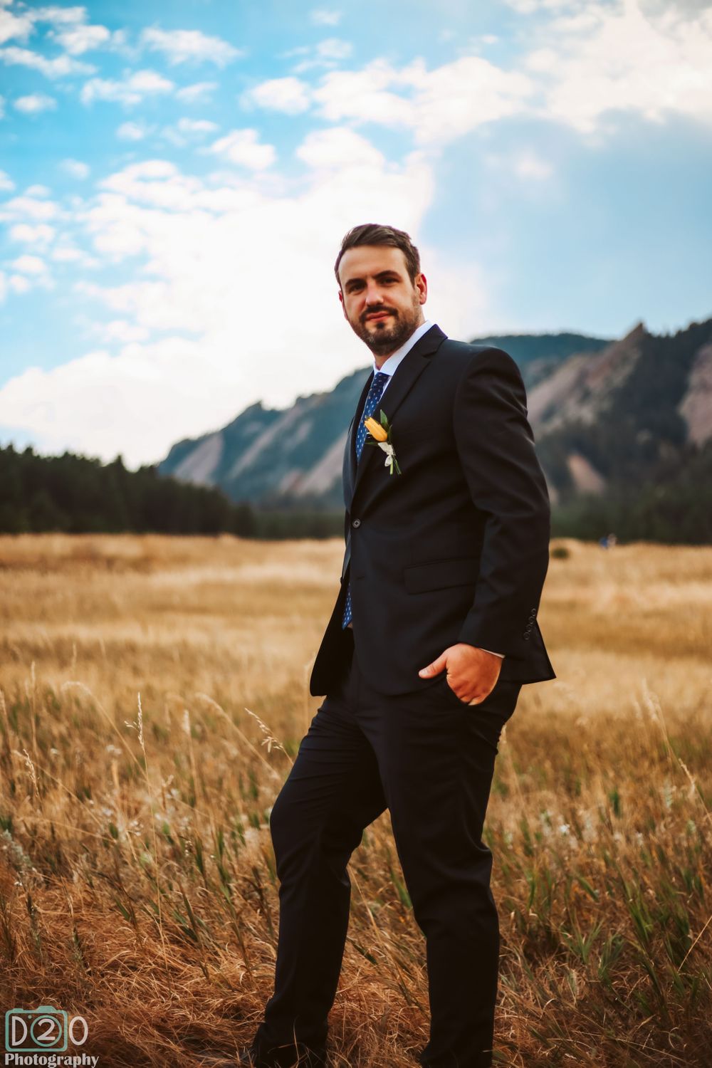 Groom in black suit poses in golden mountain meadow on wedding day.