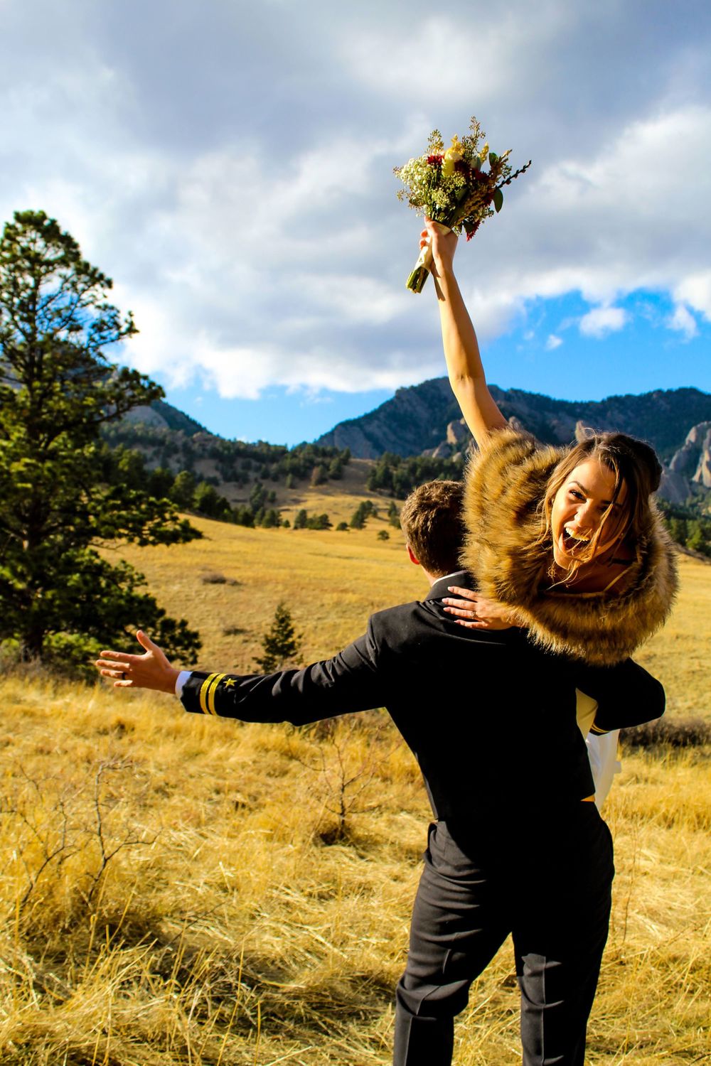 Joyful couple embracing in mountain field with bouquet raised triumphantly.