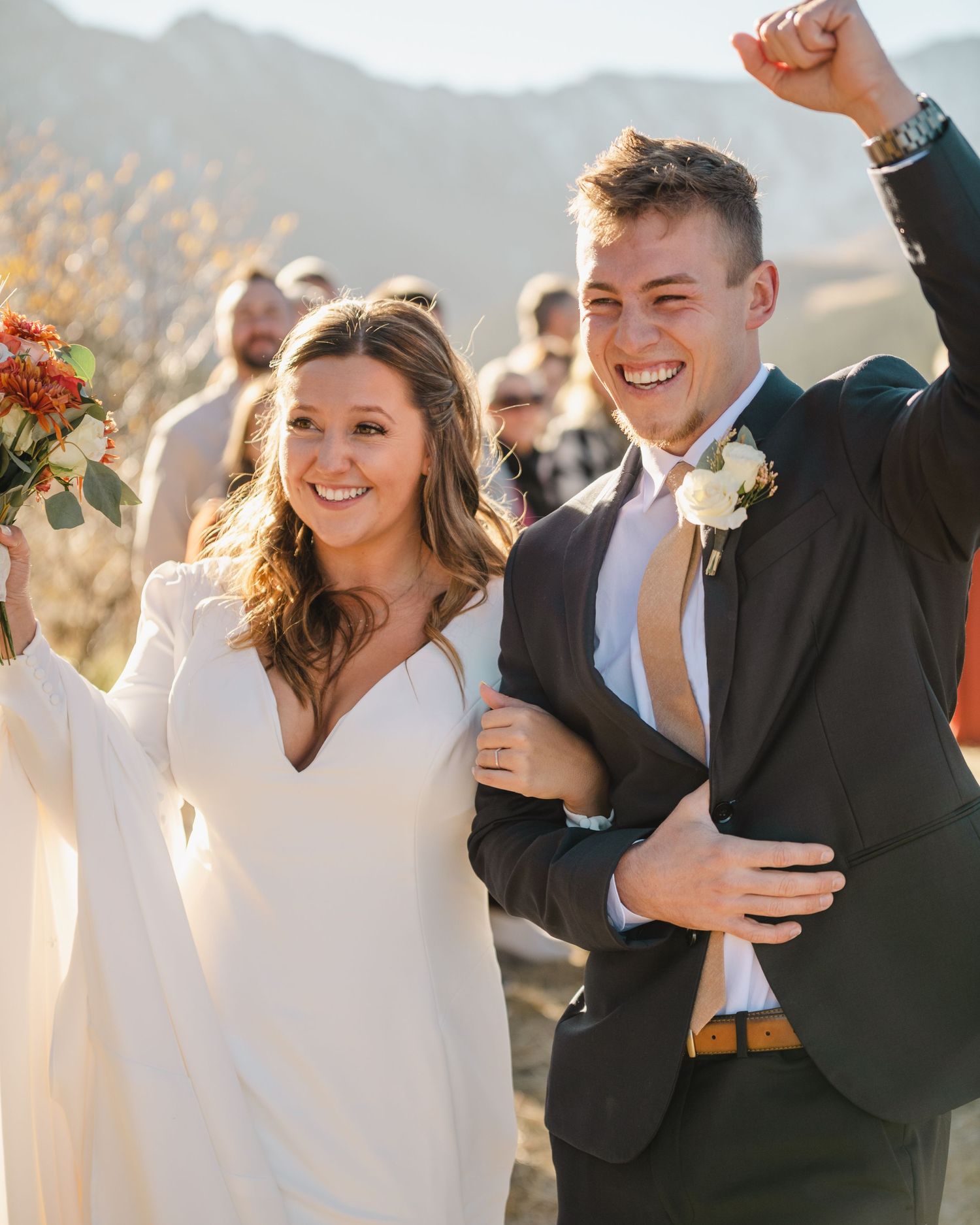 Wedding couple celebrates joyfully with arms raised during outdoor mountain ceremony with fall colors in background.