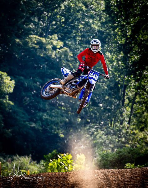 Motocross rider performing dramatic jump through sunlit forest setting.