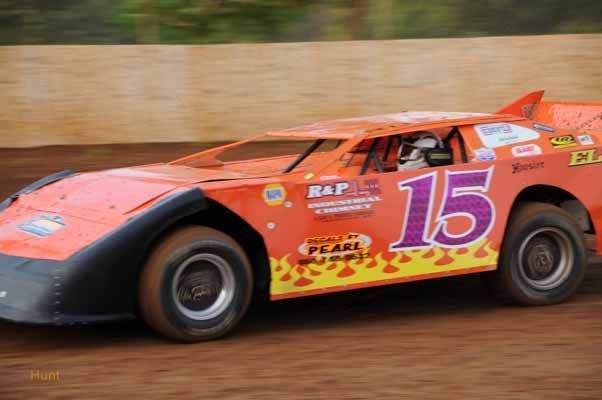 Red race car number 15 speeding along dirt track during competitive motorsport event.