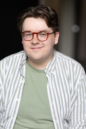 Professional headshot of a person wearing striped shirt and green t-shirt with red glasses.