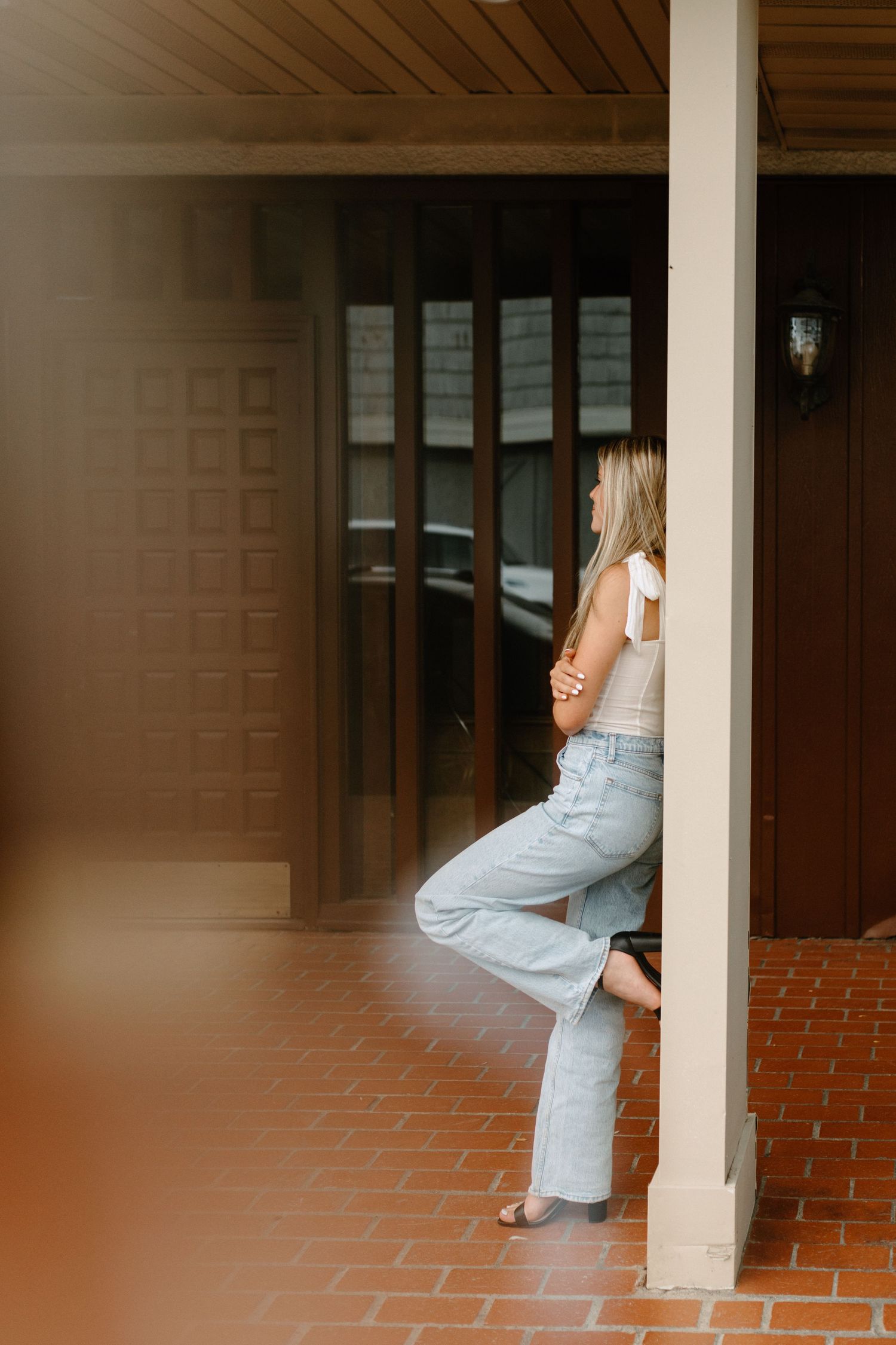 Person in casual denim leans against white column on brick patio in soft lighting.