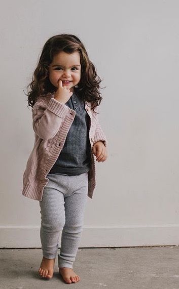 Toddler wearing pink sweater, grey pants and dark vest against grey background.