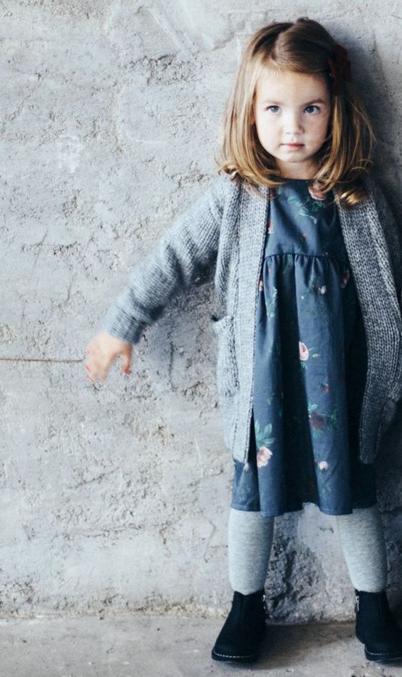 Young child in gray cardigan and blue dress stands against textured wall in stylish winter outfit.