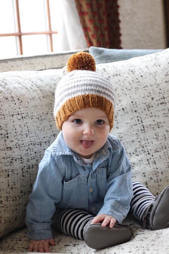 Baby wearing pom pom beanie and denim shirt crawls on gray couch.