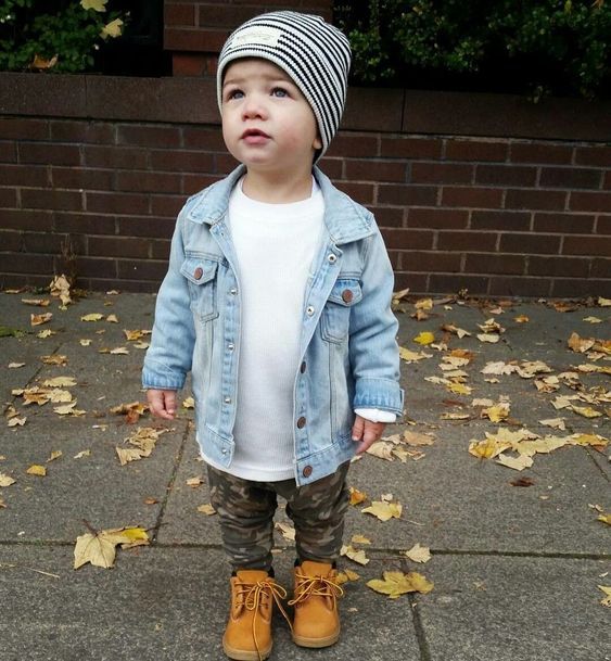 Small child in denim jacket and striped beanie standing on sidewalk with autumn leaves.