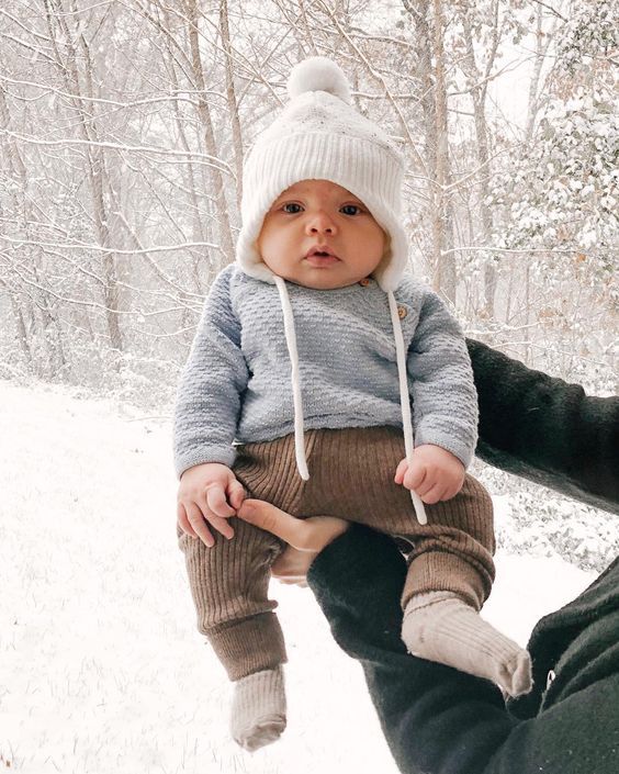 Baby in white knit hat and gray sweater sits on lap against snowy winter background.