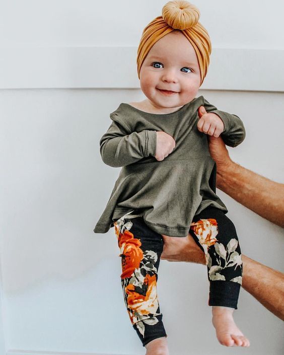 Adorable baby wearing olive green top and floral leggings with mustard headwrap sitting on someone's hands.