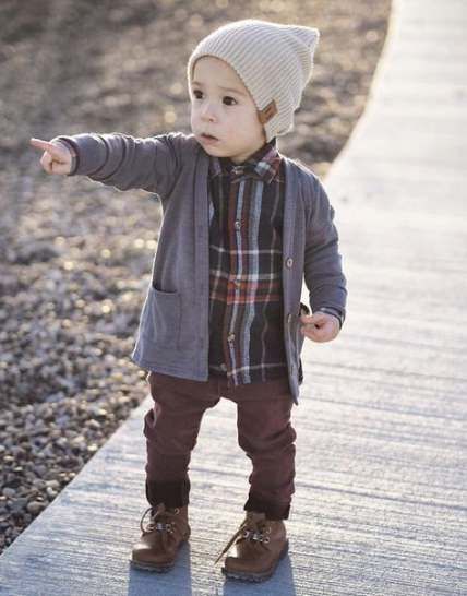 Toddler in beanie hat, plaid shirt and gray jacket points while standing on a path outdoors.