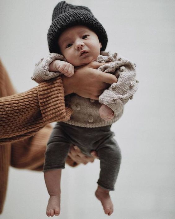 Baby wearing knit sweater and gray pants held by person in orange sleeve.