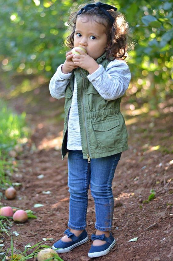 Child in olive green vest and jeans eating apple in orchard setting.