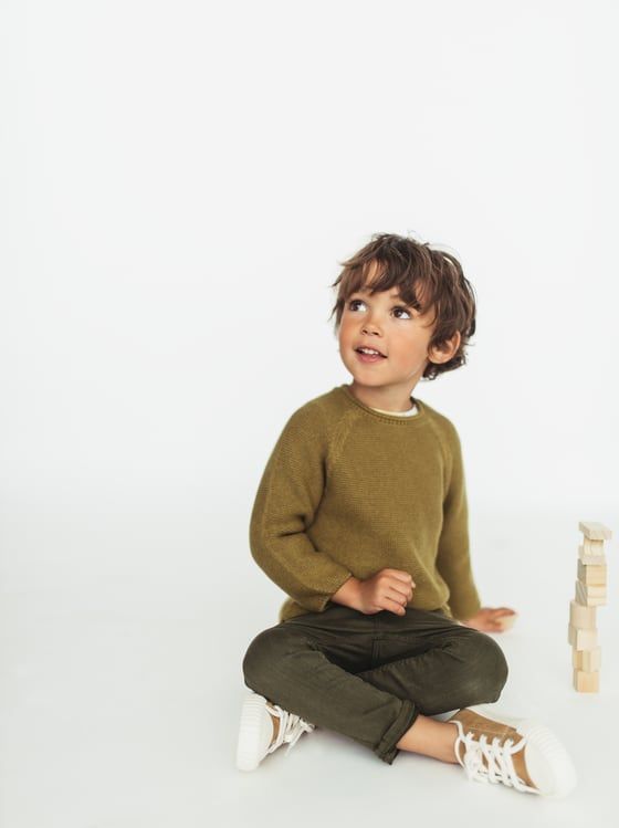 Child in olive sweater and dark pants sits cross-legged against white background.
